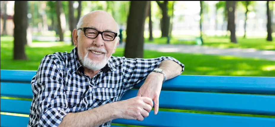 senior male in checkered blue buttonup with arm resting on bench smiling in park
