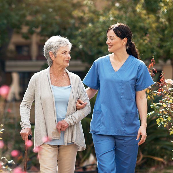 older senior woman with cane walking with nurse dressed in blue scrubs smiling with arms interlocked