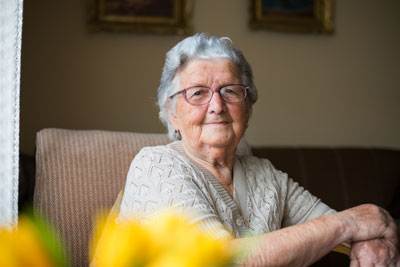 senior female sitting in chair by window wearing white sweater and smiling while looking at yellow roses