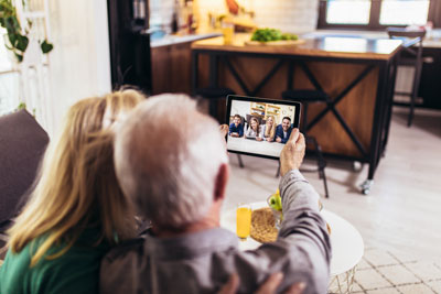 senior male and senior female sitting on couch of apartment holding iPad and video chatting with family